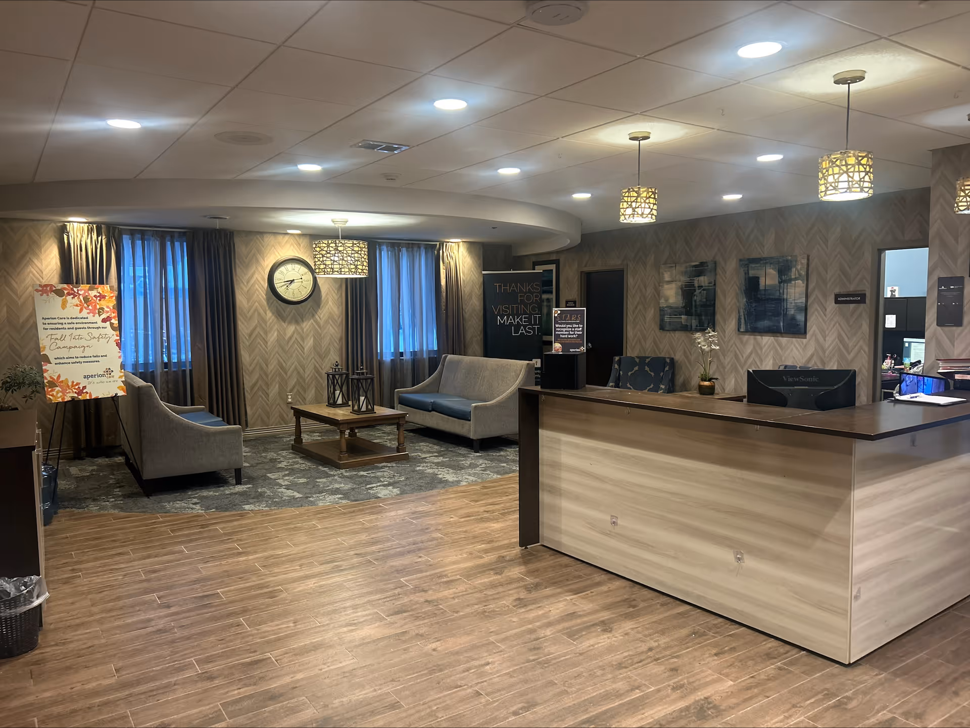 A reception area in a senior living facility with a wooden front desk on the right, two gray sofas and a wooden coffee table with lanterns in the seating area on the left. The room has wood flooring, patterned wallpaper, and three pendant lights hanging from the ceiling. There are two windows with curtains, a wall clock, and framed artwork on the walls. A sign on an easel and a thank you banner are also visible.
