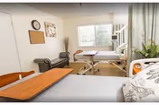 A bright and clean bedroom in a senior living facility with two hospital-style beds, a window letting in natural light, a small bench, wall decorations including a clock and a corkboard, and a plant near the window.