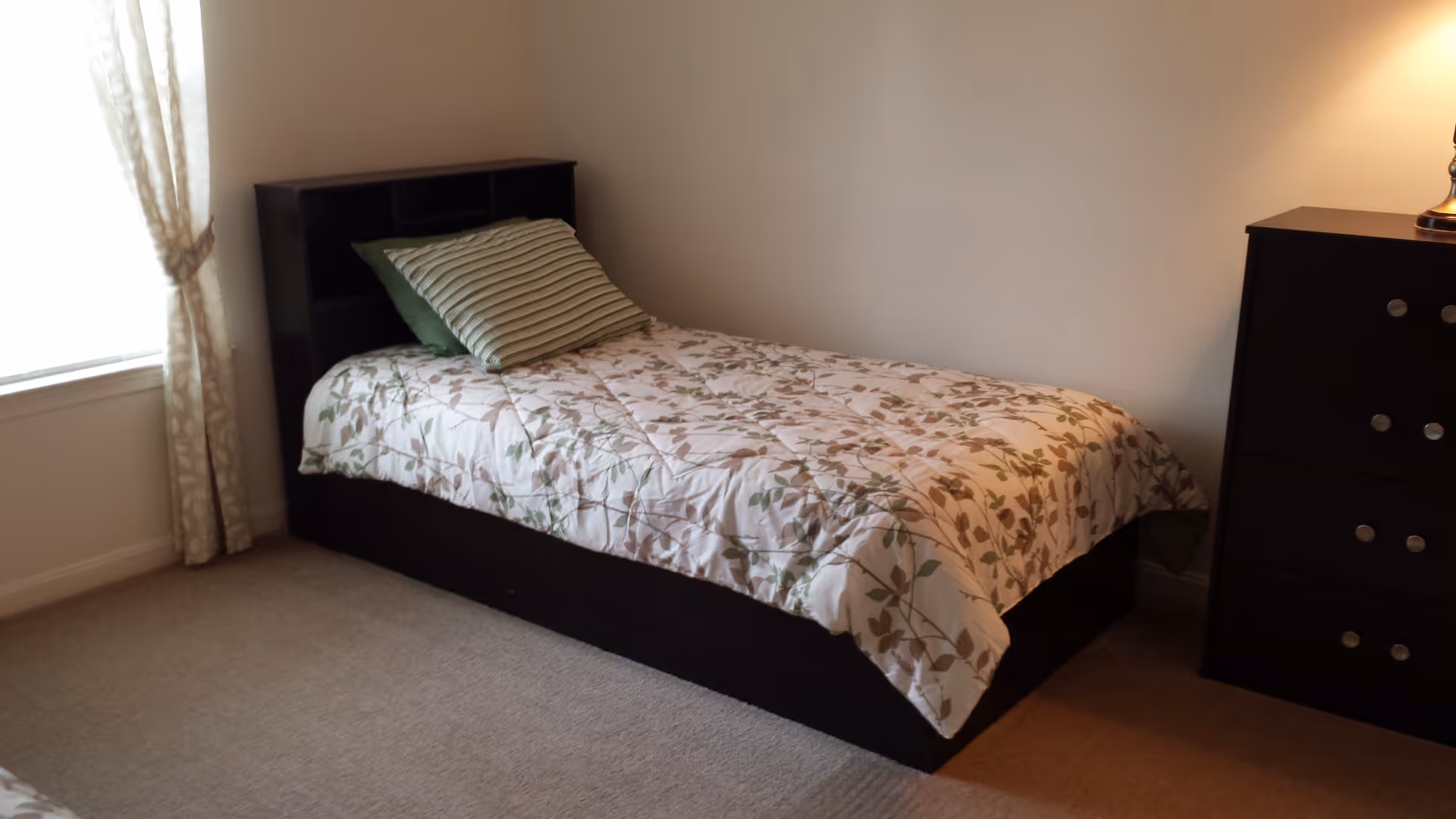 A simple bedroom with a single bed featuring a floral patterned comforter and two pillows, one striped and one solid green. The bed has a dark wooden headboard with shelves. Next to the bed is a dark wooden dresser with a lamp on top. A window with light-colored curtains is on the left side, allowing natural light to brighten the room.