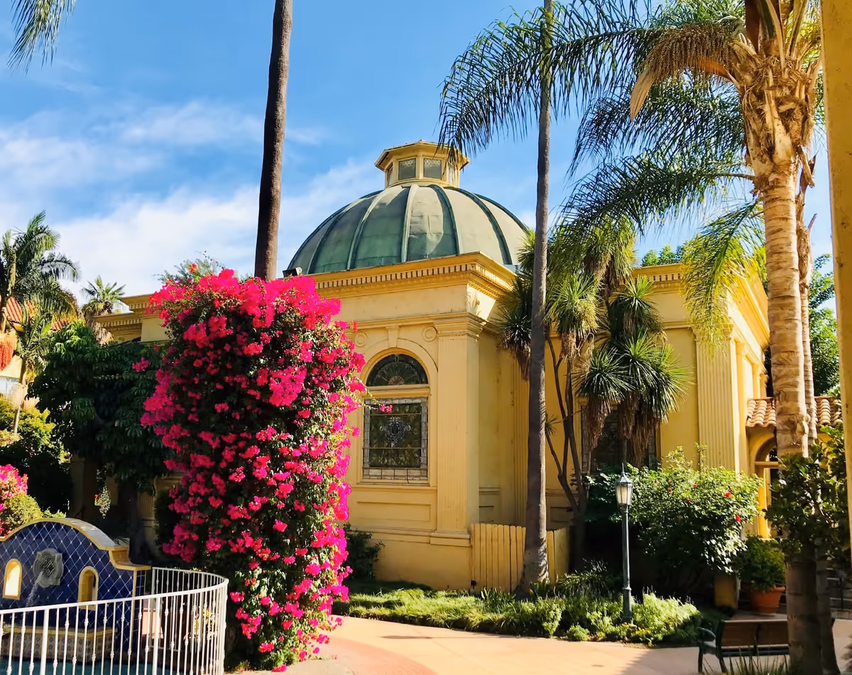 Exterior view of a yellow building with a green domed roof surrounded by palm trees and vibrant pink flowering bushes under a clear blue sky.