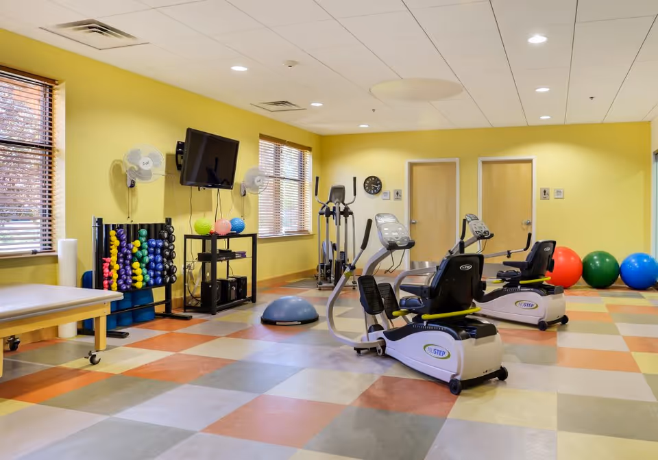 A brightly lit exercise room with yellow walls and multicolored tiled floor. The room contains two recumbent exercise bikes, an elliptical machine, a rack of colorful dumbbells, exercise balls, a balance trainer, and a wall-mounted TV. There are two windows with blinds and two closed doors on the far wall.