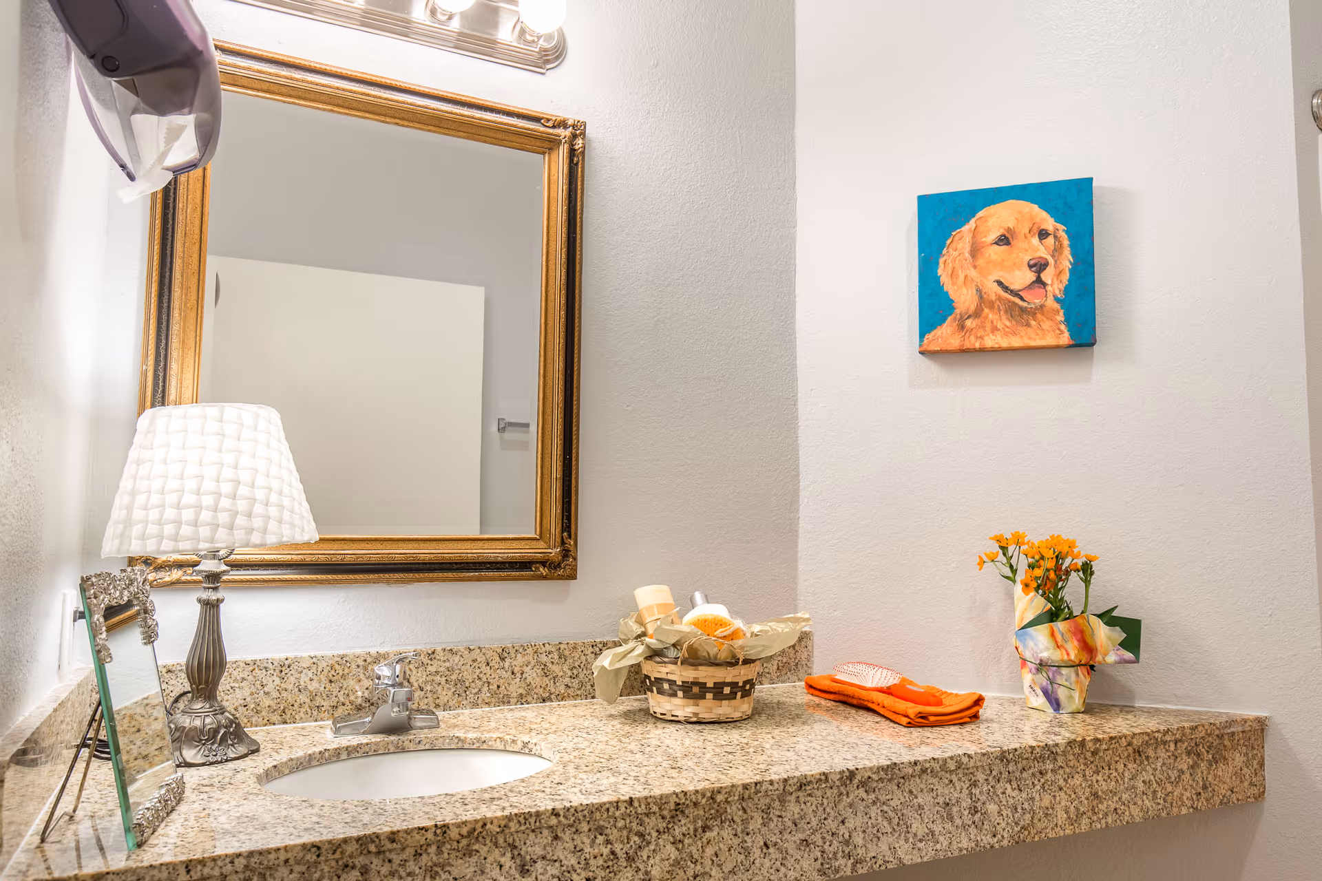 Granite bathroom vanity with a sink, gold-framed mirror, lamp, decorative basket and towels, and a small dog painting on the wall.