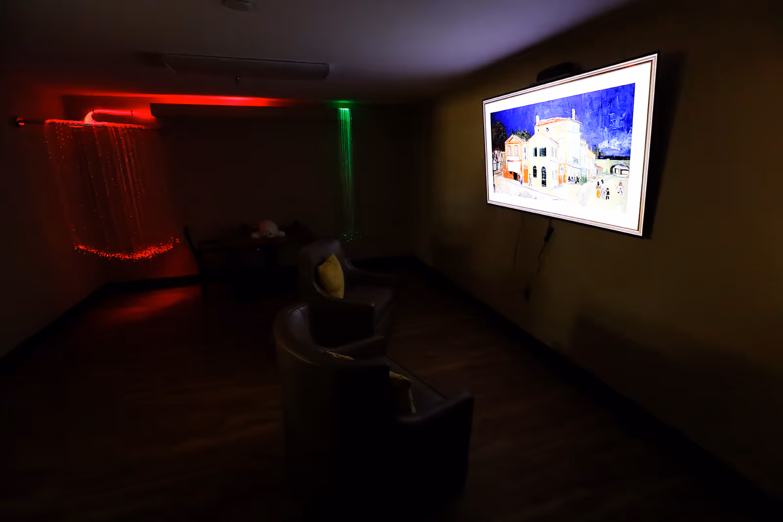 A dimly lit room with two armchairs facing a wall-mounted screen displaying a painting of buildings under a blue sky. In the corner, there are red and green fiber optic lights hanging from the ceiling, creating a calming ambiance. A small table with chairs is also visible in the background.