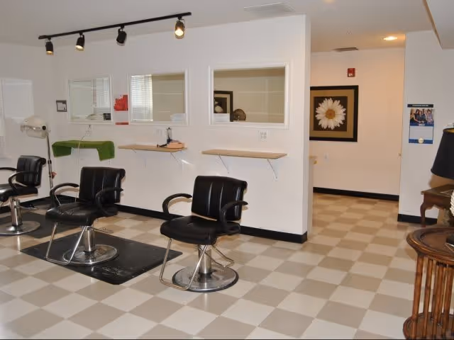Interior view of a senior community salon area with three black salon chairs positioned in front of a wall with three windows and small wooden shelves. The floor has a checkered pattern, and there is a framed flower picture on the wall in the hallway.