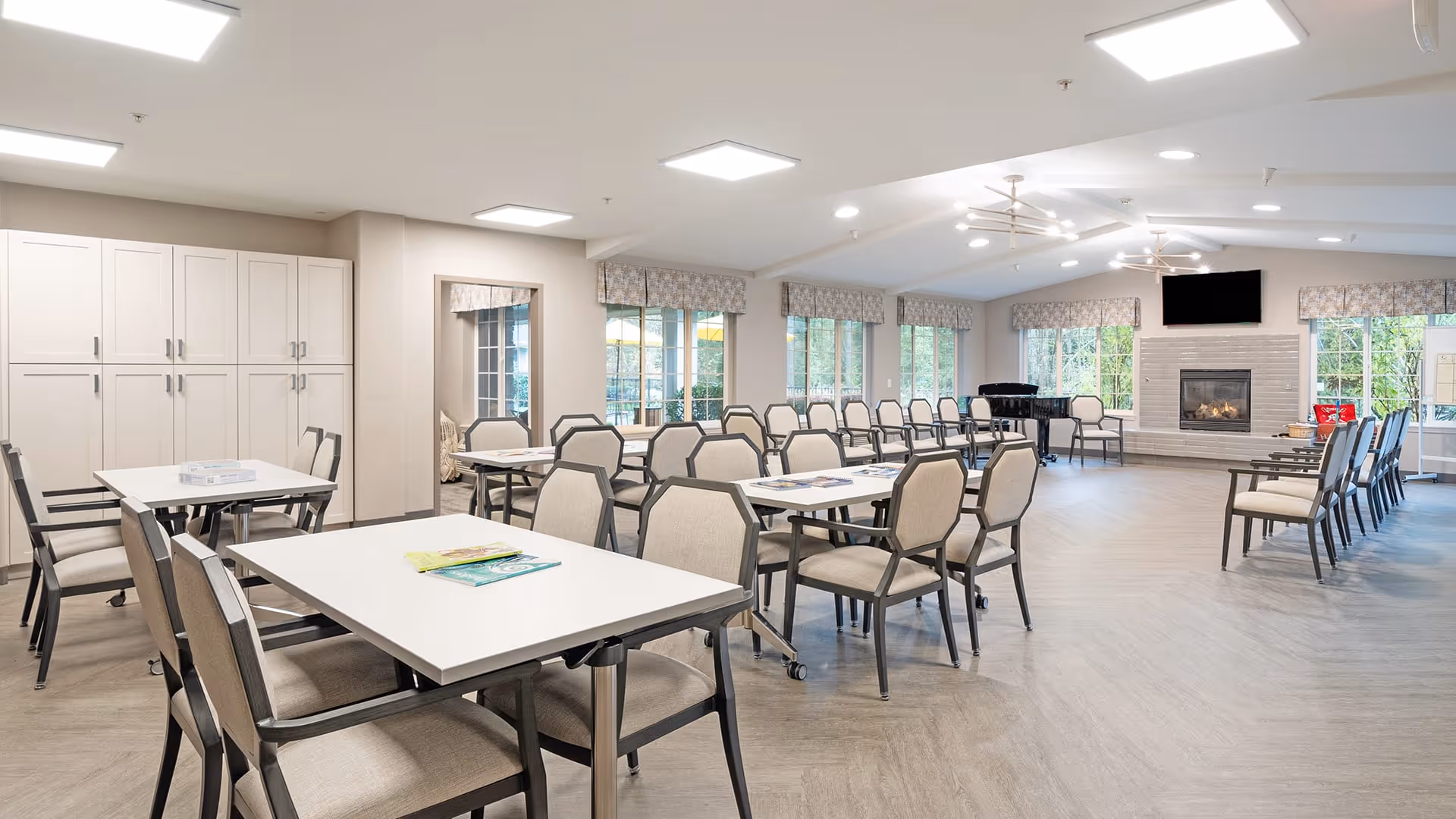 A spacious, well-lit common area with multiple tables and chairs arranged for group seating. The room features large windows with patterned valances, a fireplace with a mounted TV above it, and a piano in the corner. The floor is light-colored wood, and the ceiling has modern light fixtures.