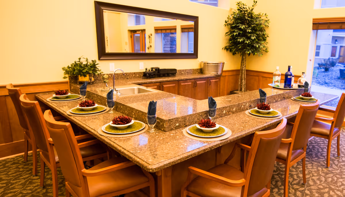 A well-lit kitchen area with a large granite countertop island surrounded by wooden chairs with leather seats. The countertop is set with plates, green placemats, bowls with red decorative flowers, and glasses with folded dark napkins. There is a sink in the countertop, a large mirror on the wall, a potted plant in the corner, and bottles of liquor on a tray near a window showing an outdoor view.