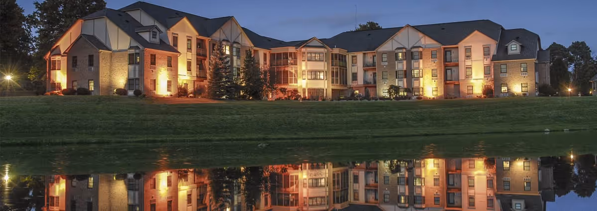 A multi-story senior living building lit at dusk with its lights reflected in a calm pond.