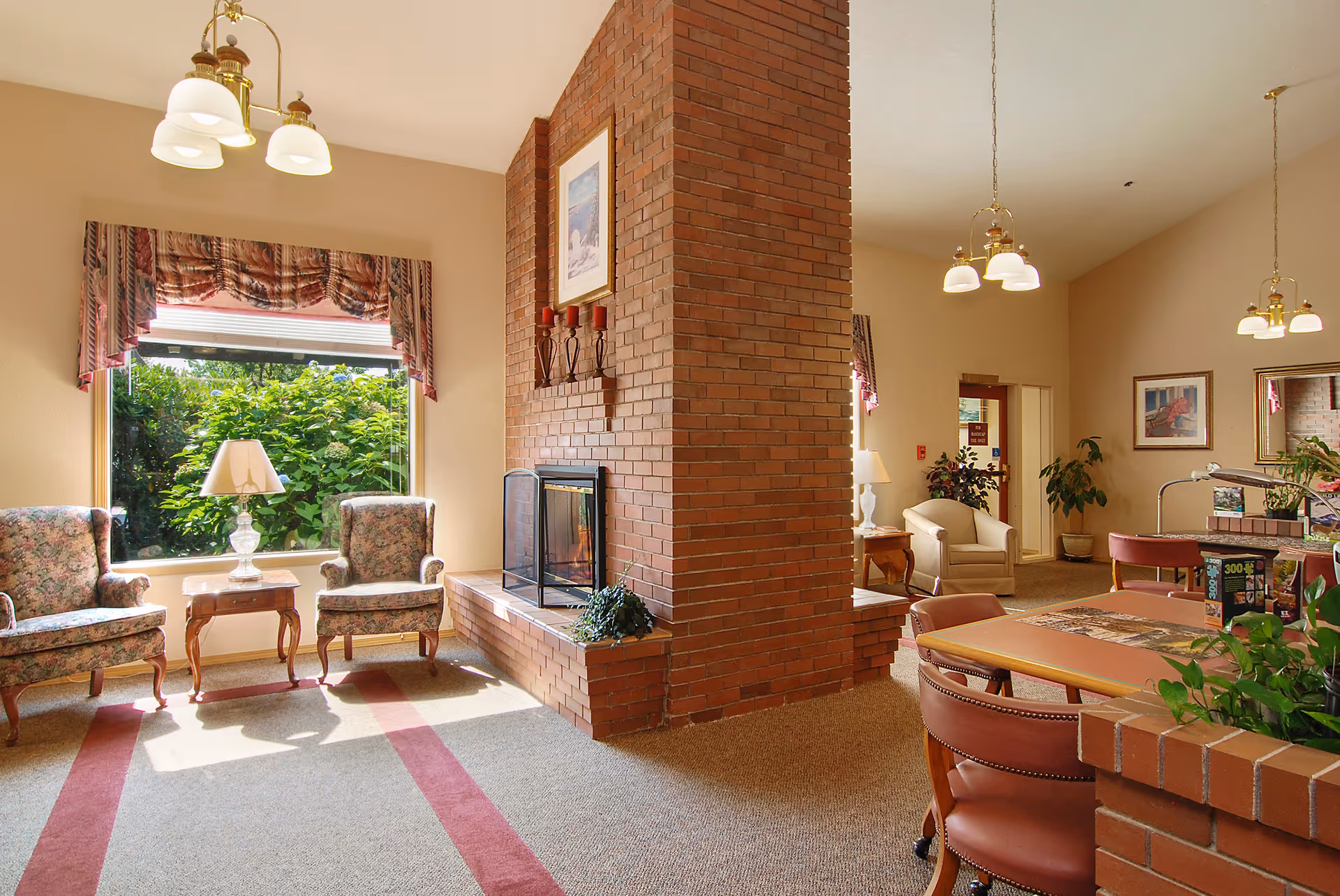 A cozy common area with a large brick fireplace in the center. Two floral armchairs and a small wooden table with a lamp are positioned near a window with patterned valances, letting in natural light. On the other side of the fireplace, there are additional seating areas with chairs and tables, plants, framed artwork on the walls, and hanging light fixtures.
