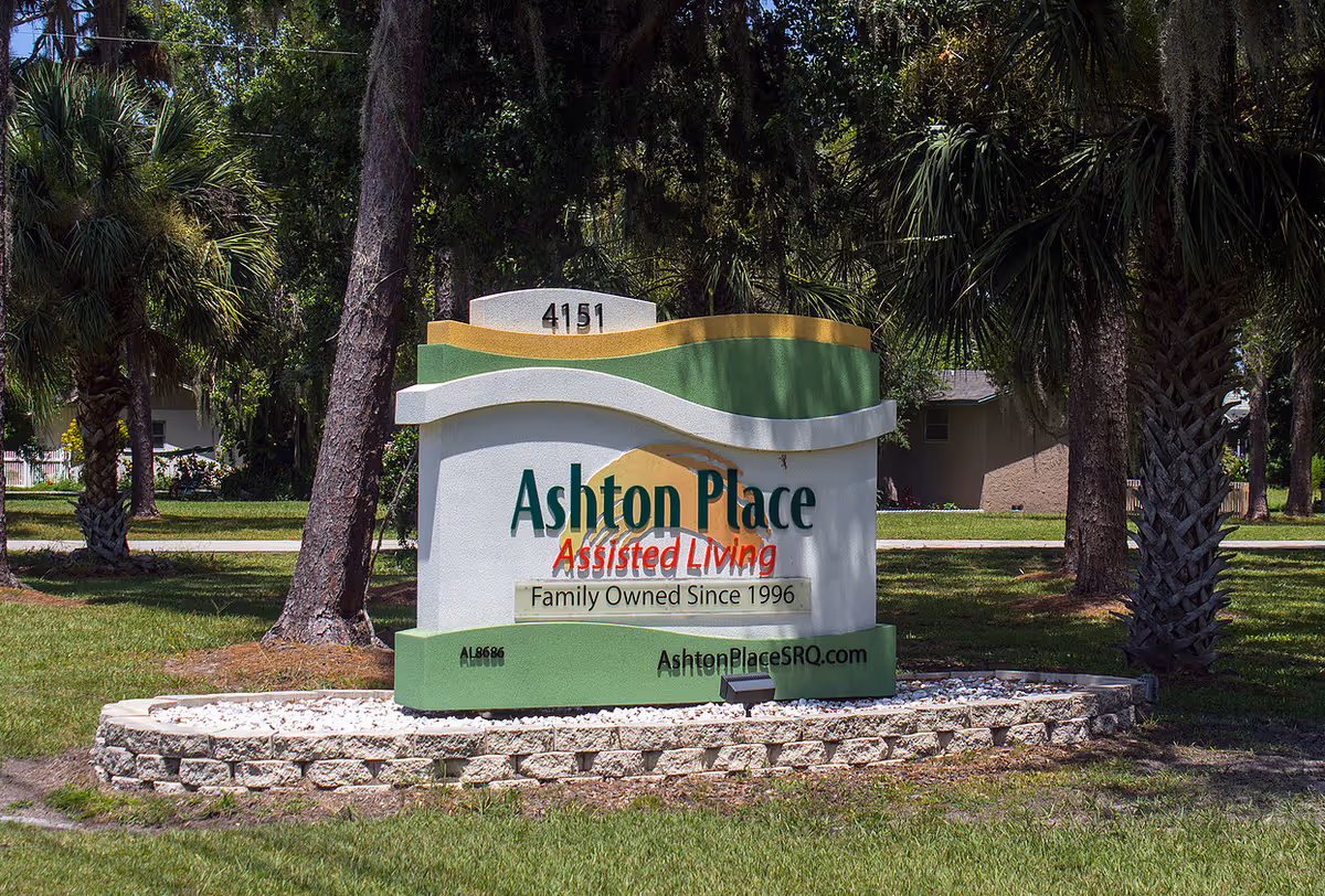 Outdoor view of a large sign for Ashton Place Assisted Living, surrounded by trees and grass. The sign includes the address number 4151, the phrase 'Family Owned Since 1996', and the website AshtonPlaceSRQ.com.