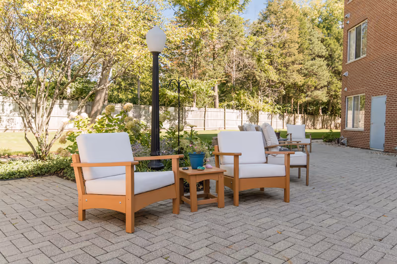 Outdoor patio area with cushioned wooden chairs and a small wooden table on a paved surface, surrounded by greenery and trees, next to a brick building.