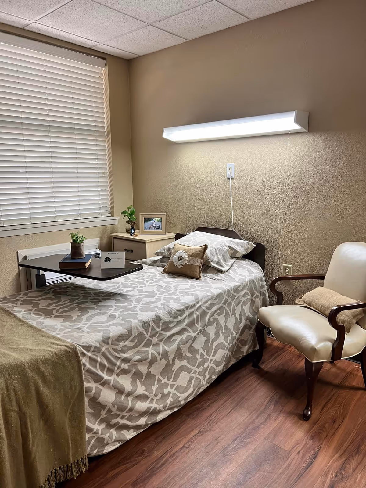 A tidy nursing home bedroom with a single bed, bedside table and overbed tray, upholstered chair, window with blinds and a wall-mounted light.