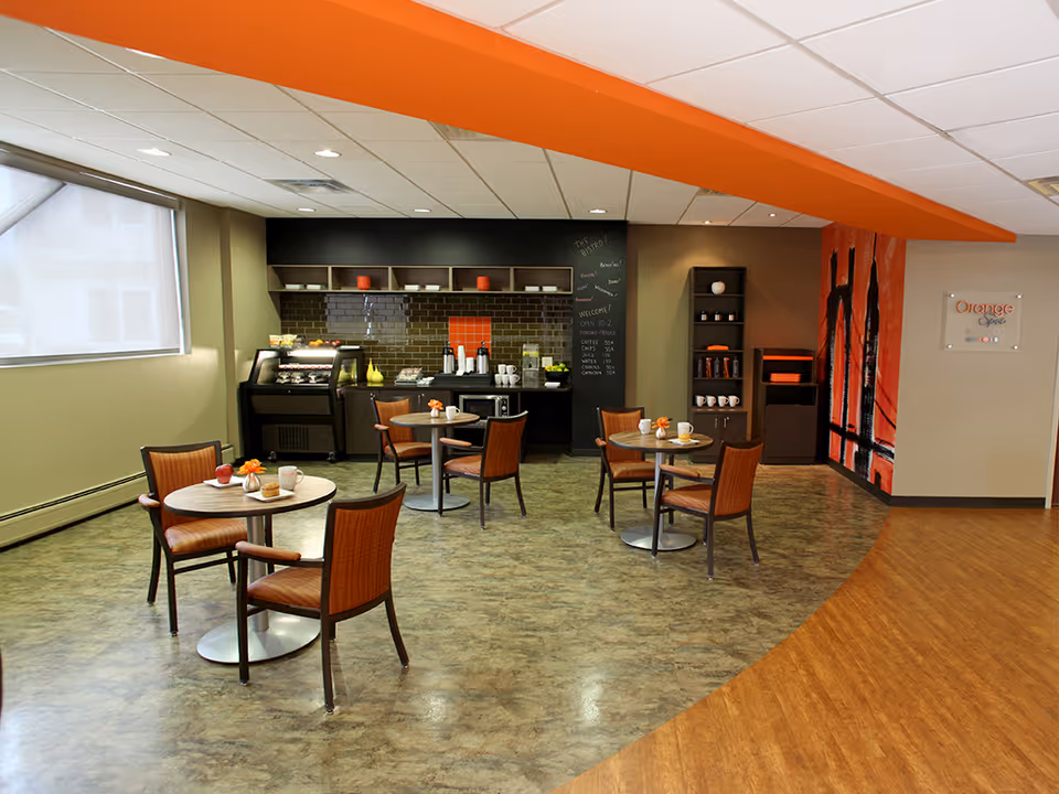 A small dining area with four round tables, each surrounded by two wooden chairs with orange cushions. The room has a coffee and snack station against the back wall with shelves, a chalkboard menu, and a small refrigerator. The floor is a mix of wood and tile, and the ceiling has recessed lighting with an orange beam running across it. A large window with a roller shade is on the left side, and a decorative orange and black wall art is on the right.