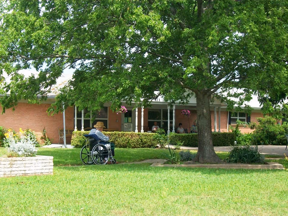 A person in a wheelchair sits on the lawn under a large tree in front of a brick nursing facility with a covered porch.