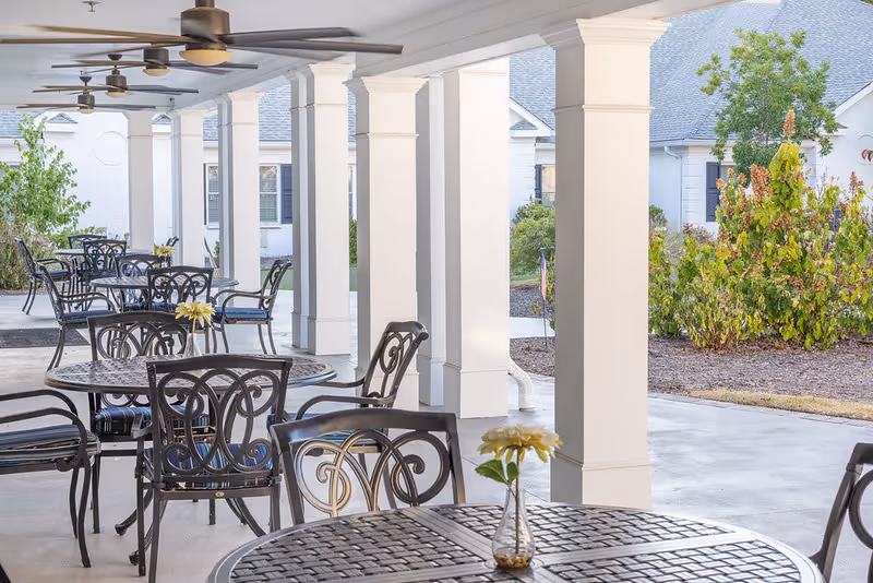 Covered outdoor patio with multiple metal tables and chairs and ceiling fans overlooking landscaped grounds.