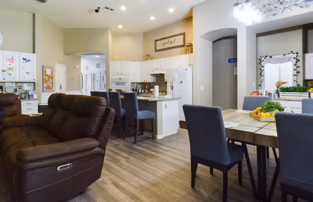 Interior view of a senior living facility showing a cozy living room area with a brown recliner sofa, a kitchen with white cabinets and a breakfast bar with blue chairs, and a dining area with a wooden table and blue upholstered chairs. The space is well-lit with recessed lighting and a chandelier above the dining table. There are decorative elements including a framed 'grateful' sign and a basket of fruit on the dining table.
