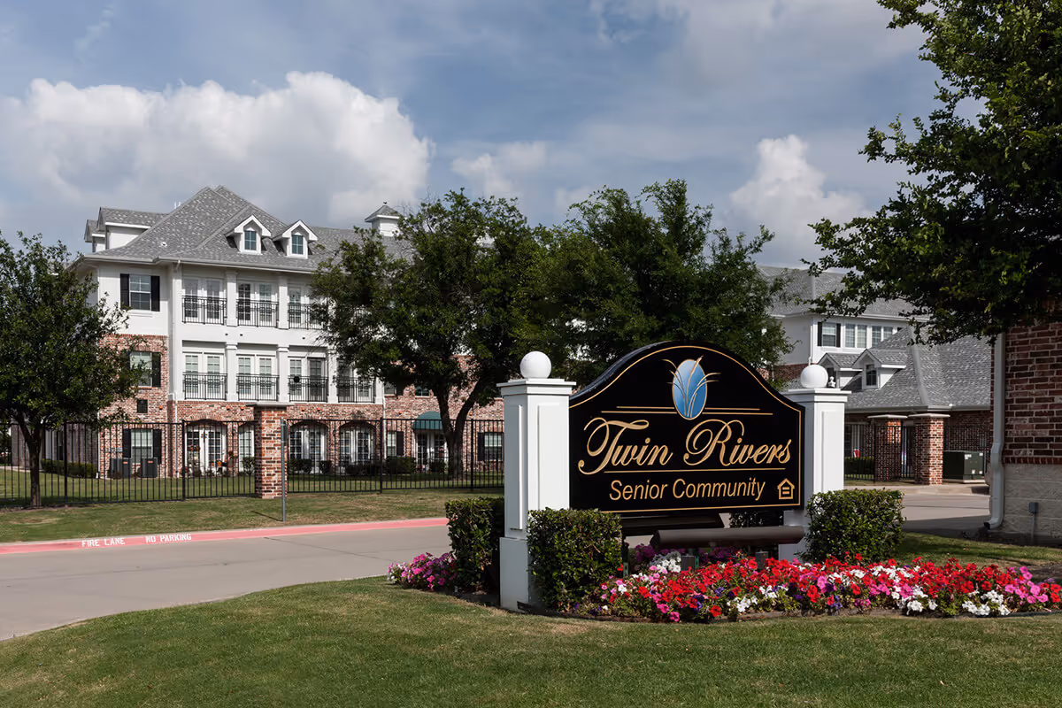 Exterior view of Twin Rivers Senior Community building with a large sign in front surrounded by colorful flowers and green grass under a partly cloudy sky.
