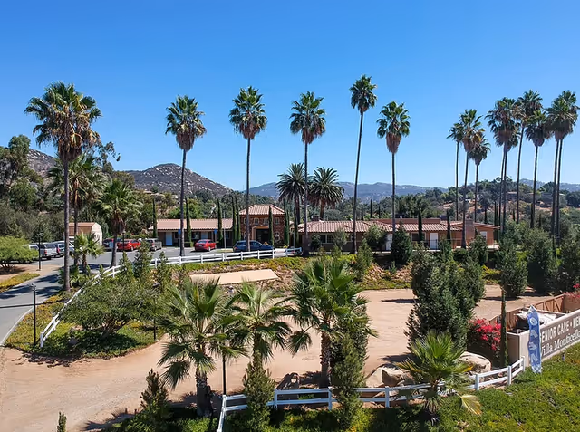 View of Villa Monticello senior living facility with a single-story building surrounded by tall palm trees, greenery, and mountains in the background under a clear blue sky. A driveway with parked cars and a sign for the facility are visible in the foreground.