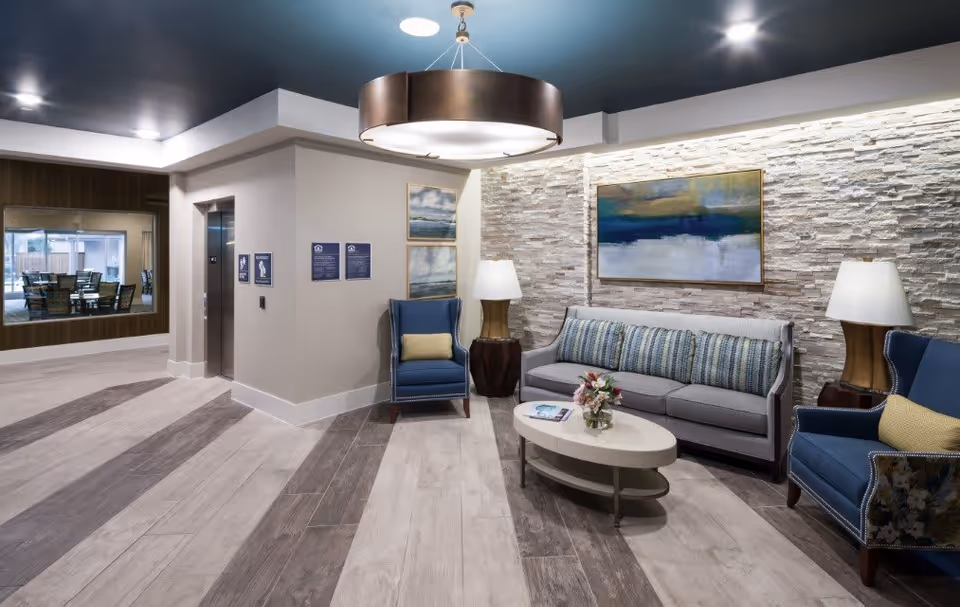 A modern and cozy seating area in a senior living facility with a gray sofa adorned with striped cushions, two blue armchairs with yellow pillows, a white oval coffee table with a small flower arrangement and a magazine, and two table lamps on dark wooden side tables. The background features a textured stone wall with a large abstract painting. To the left, there is an elevator and a window looking into a dining area with tables and chairs. The ceiling is painted blue with a large circular light fixture.