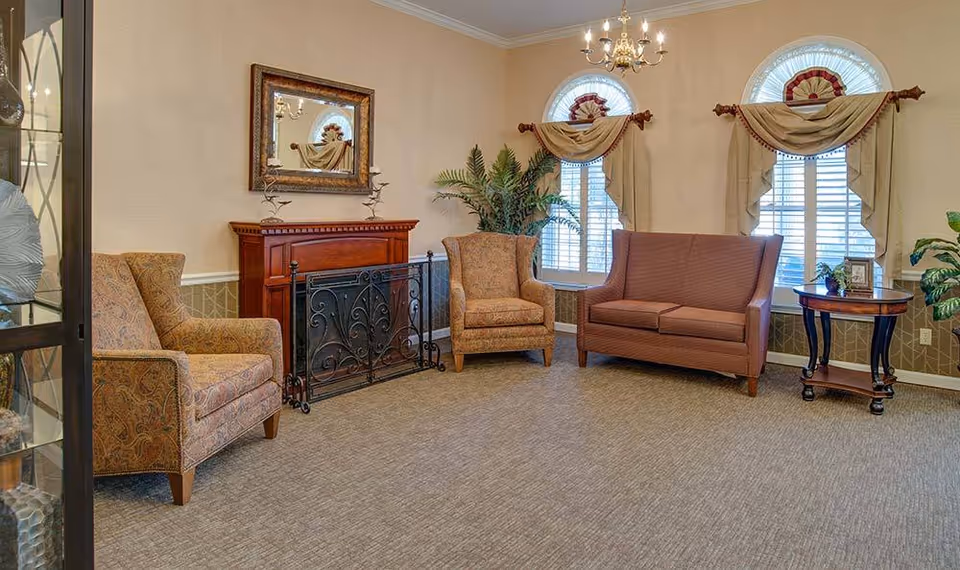 A tastefully furnished senior living lounge with upholstered chairs and a loveseat arranged around a decorative fireplace and arched windows.