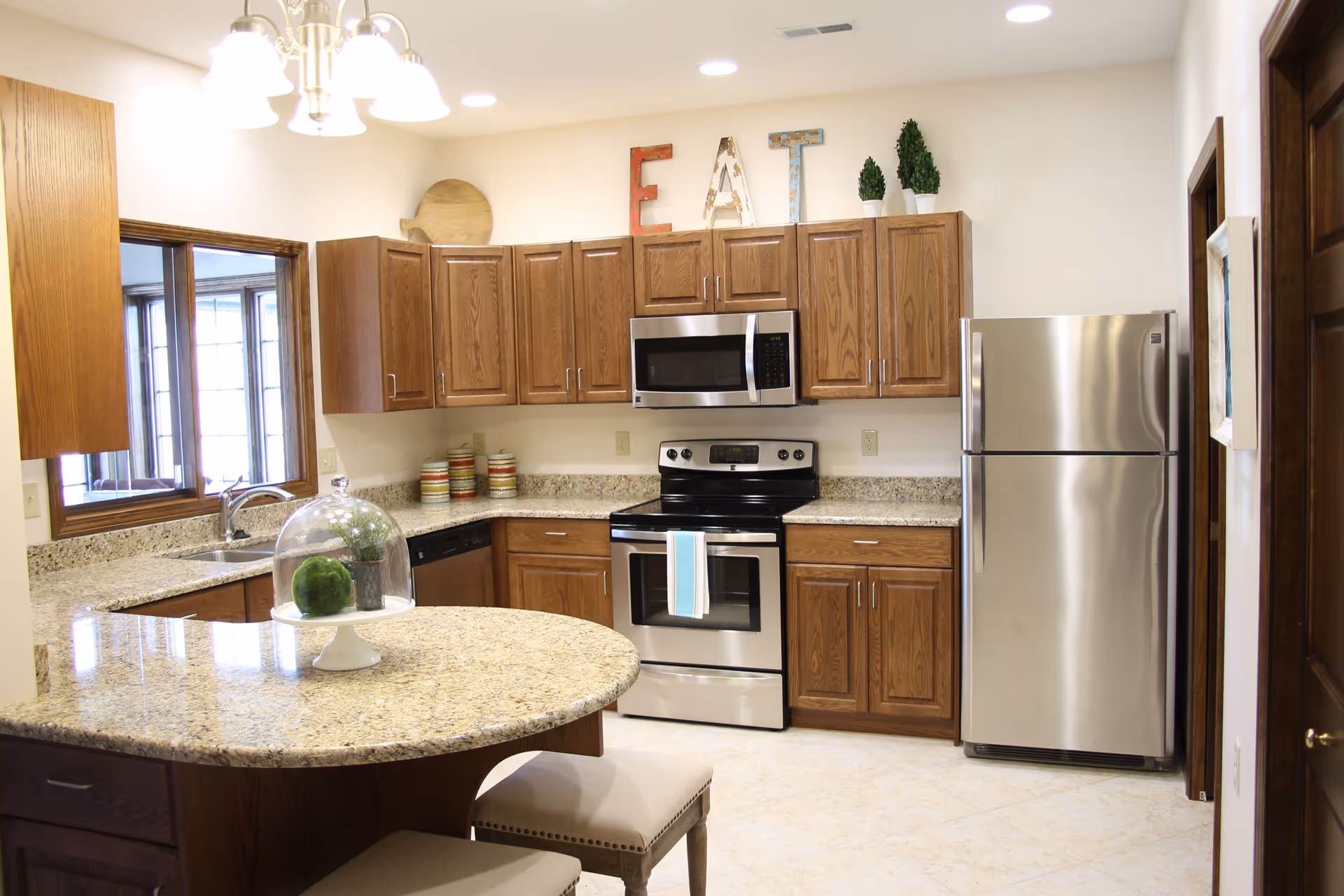 A modern kitchen with wooden cabinets, granite countertops, and stainless steel appliances including a refrigerator, stove, and microwave. There is a rounded kitchen island with two cushioned stools underneath. Above the cabinets, decorative letters spell out 'EAT' along with two small potted plants. A window is visible on the left side, allowing natural light into the space.