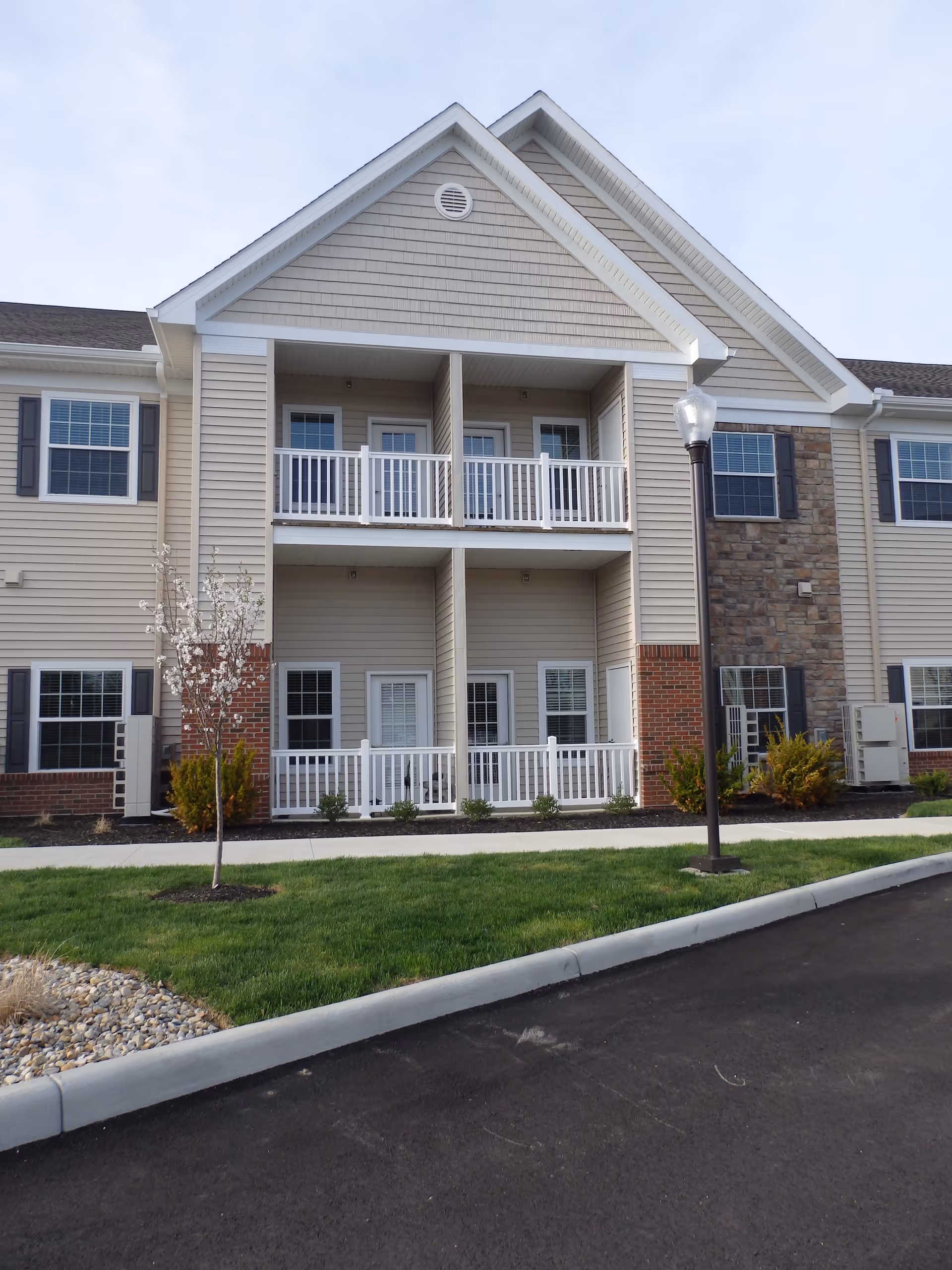 Exterior view of a two-story residential building with beige siding, white railings on balconies and patios, multiple windows with shutters, a small tree and bushes in front, a sidewalk, and a streetlamp near the curb.