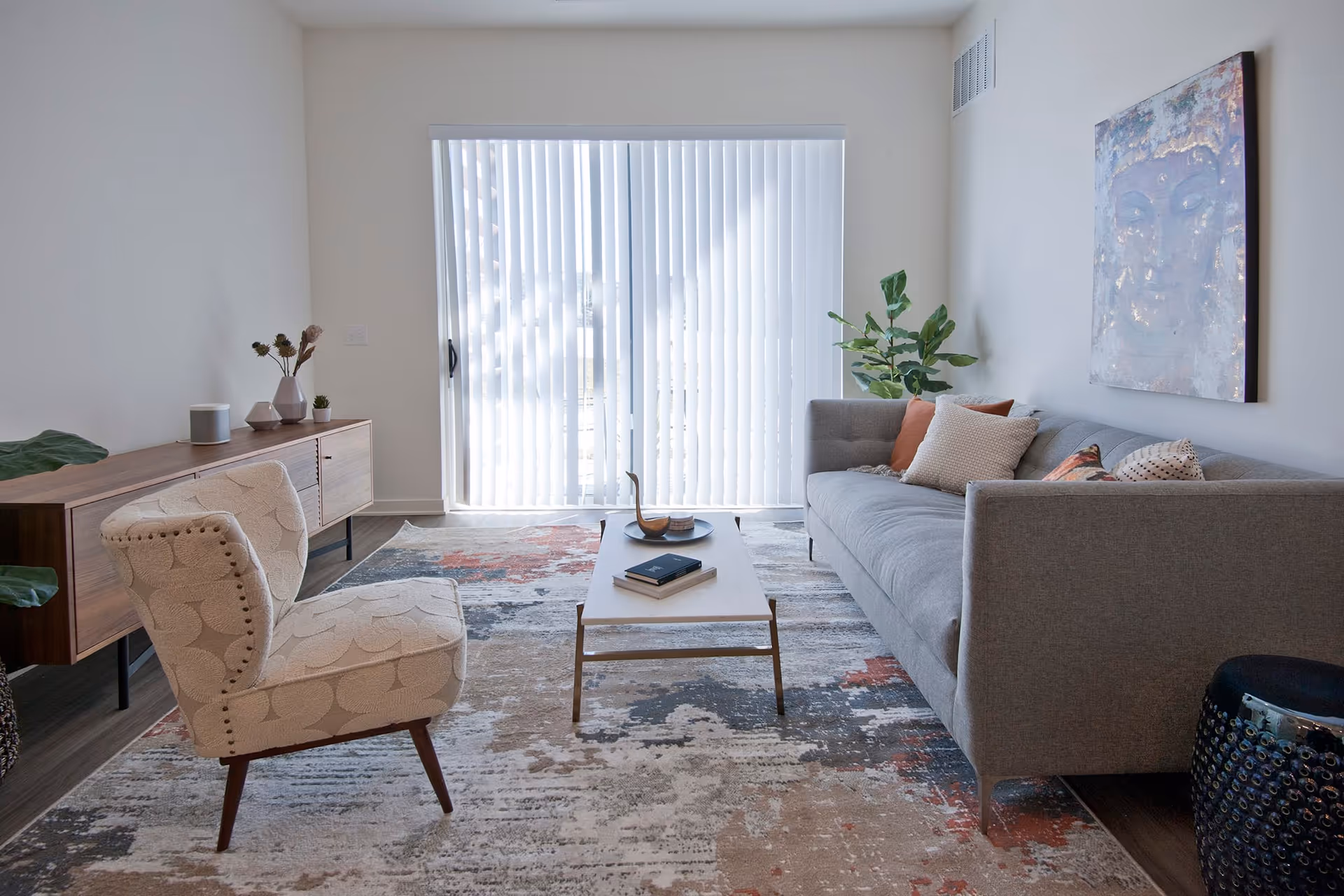 A modern living room with a gray sofa adorned with multiple pillows, a patterned armchair, a wooden coffee table with books and decorative items, a large abstract rug, a wooden sideboard with vases and plants, a large window with vertical blinds, and a large piece of artwork on the wall.