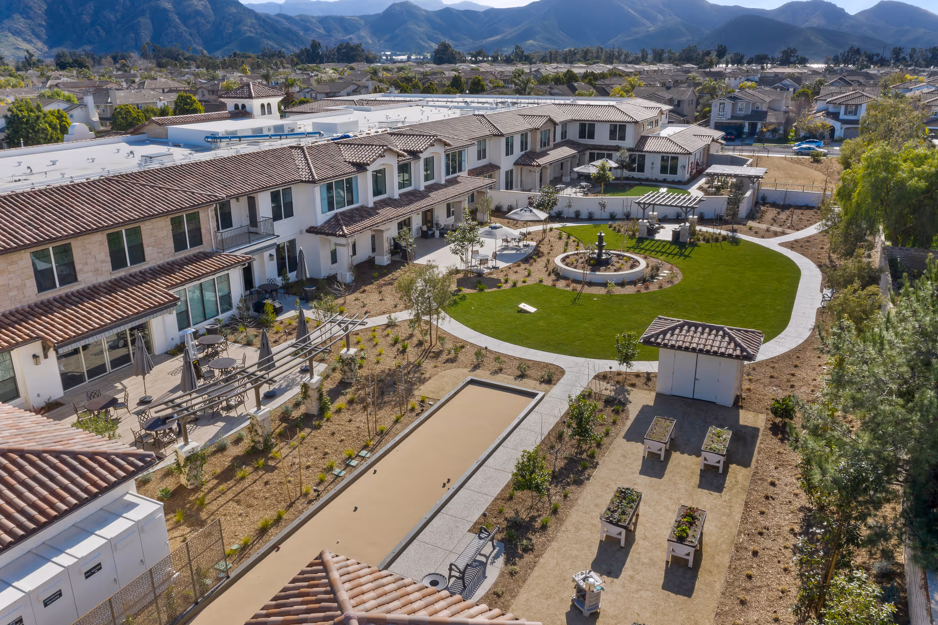 Aerial view of Oakmont of Camarillo senior living facility showing a landscaped outdoor courtyard with green lawn, walking paths, a central fountain, seating areas with tables and umbrellas, a bocce ball court, and raised garden beds. The surrounding buildings have tiled roofs and multiple windows, with mountains visible in the background.