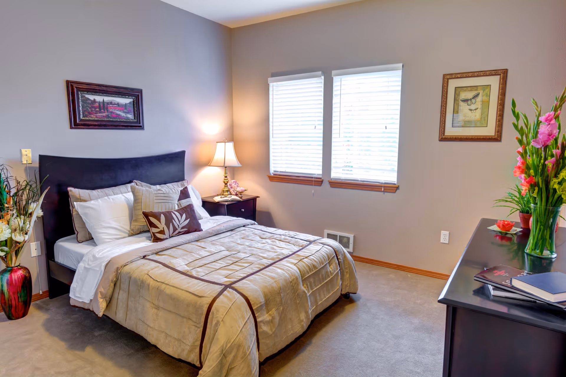 A neatly made bed with beige and brown bedding in a bedroom. The room has two windows with white blinds, a nightstand with a lamp and flowers, a dresser with books and a vase of flowers, and framed artwork on the walls. The carpet is beige and the walls are painted light gray.