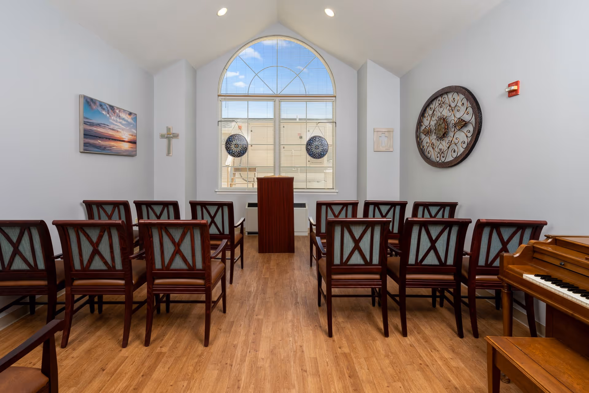 A small chapel or meditation room with wooden chairs arranged in rows facing a wooden podium. The room has a large arched window with blinds, a cross on the left wall, a decorative circular metal wall art on the right wall, and a piano in the corner. The floor is wooden and the walls are painted light gray.