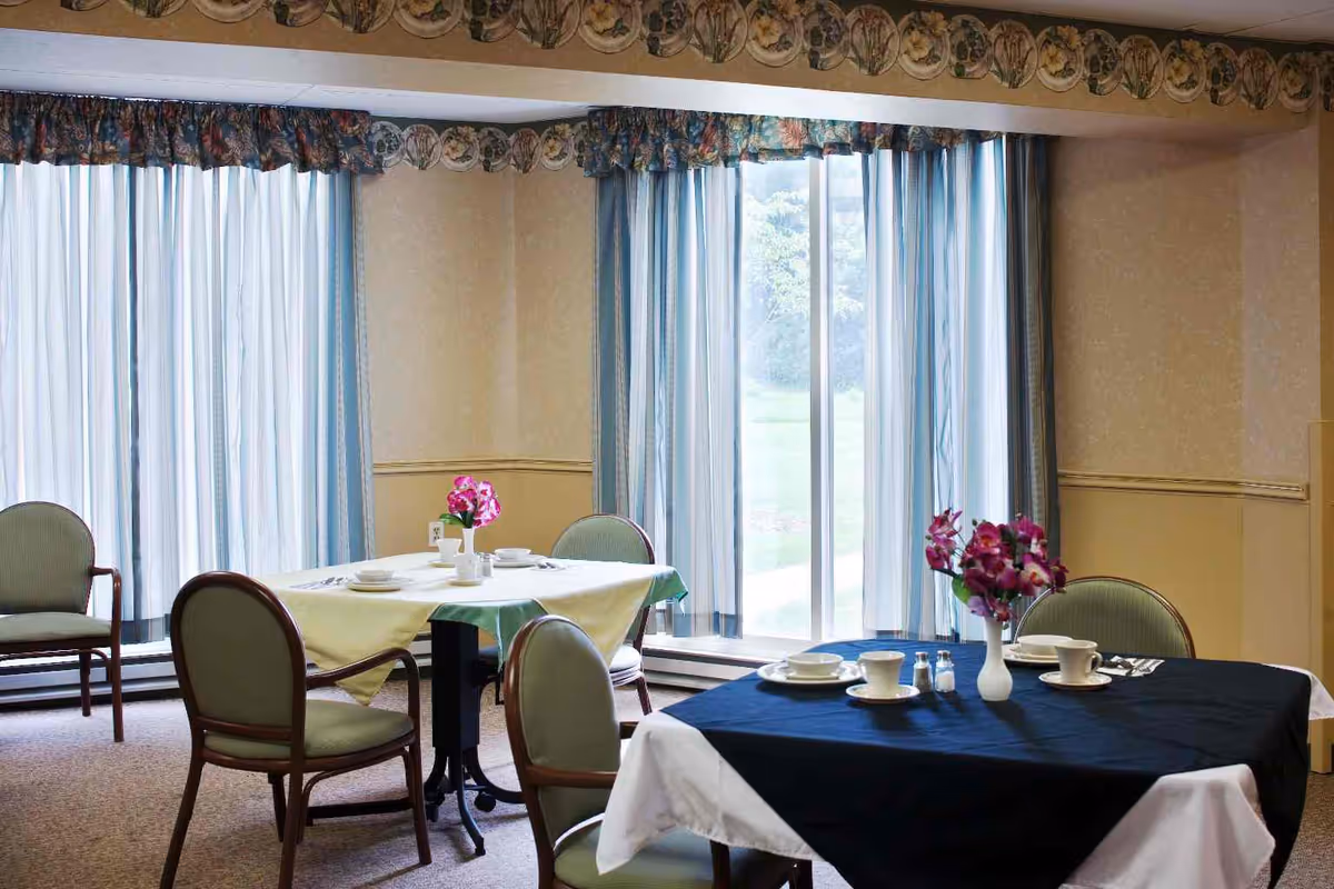 A dining room with two tables covered with tablecloths, one yellow and one black, each set with cups, plates, and a vase of purple flowers. The room has large windows with blue and white curtains and several green cushioned chairs around the tables.