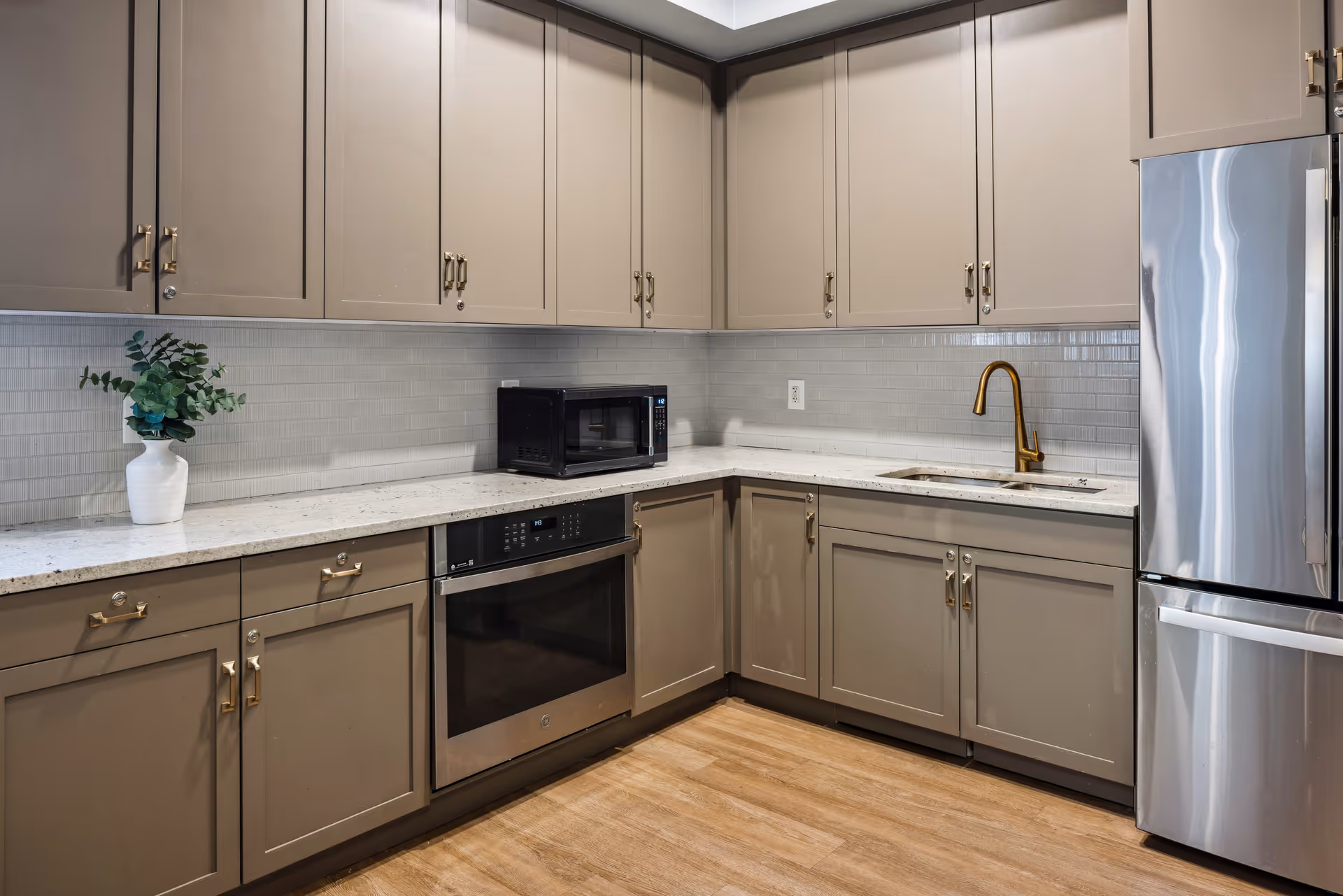 Modern kitchen with beige cabinets, a white countertop, a built-in oven, a microwave, a stainless steel refrigerator, a gold faucet, and a white vase with green foliage on the counter.