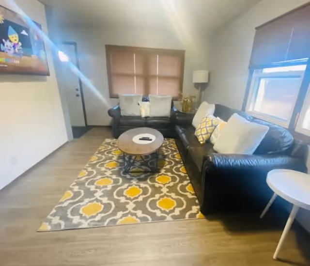 Cozy living room with two black leather sofas arranged around a wooden oval coffee table on a gray and yellow patterned rug, with windows and blinds.