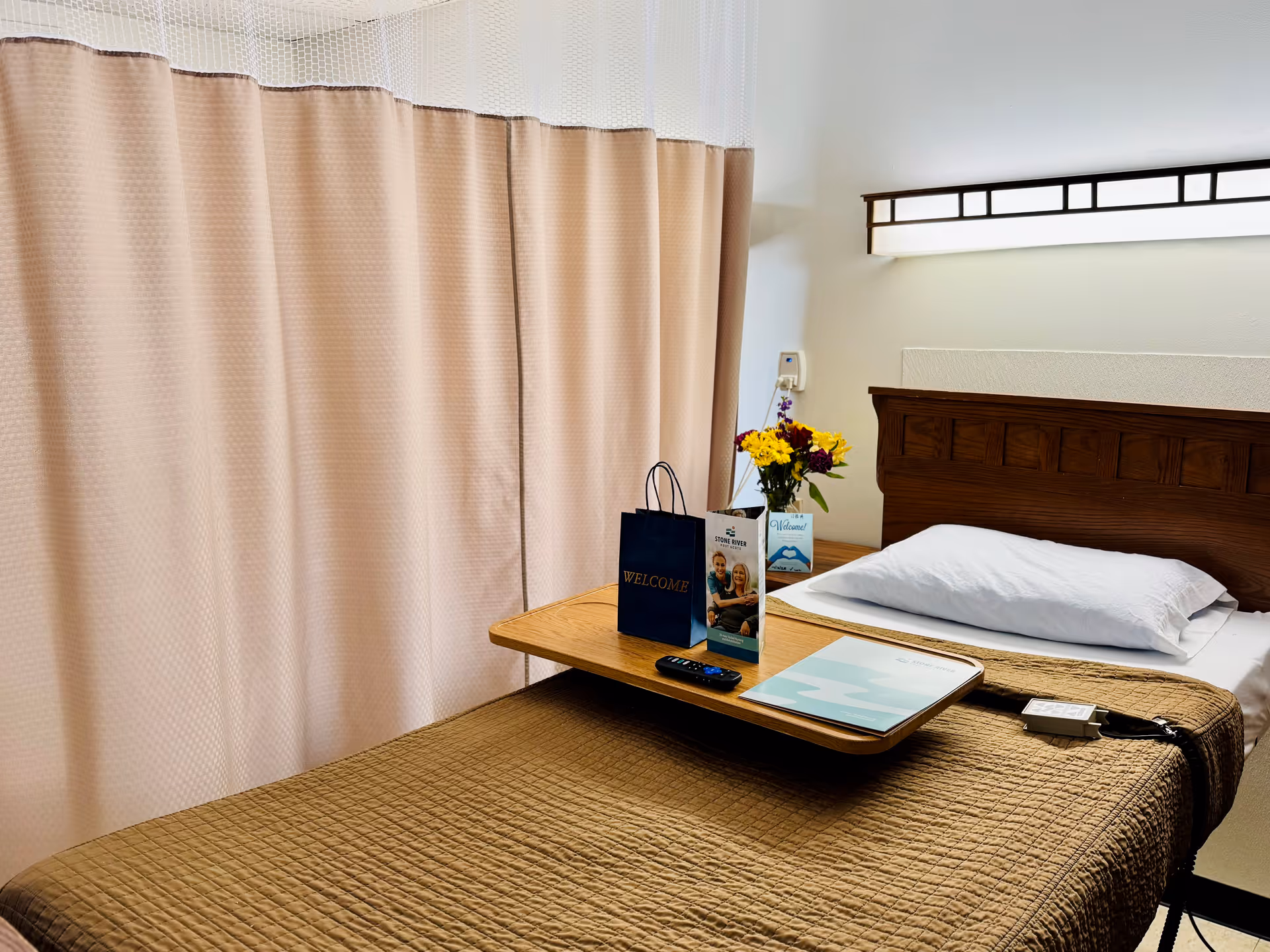 A single bed in a room with a brown quilted blanket and a white pillow. A wooden headboard is visible, along with a wall-mounted light fixture above it. A wooden overbed table holds a remote control, a welcome bag, a brochure, a small vase with yellow and purple flowers, and a welcome card. Beige privacy curtains are drawn in the background.