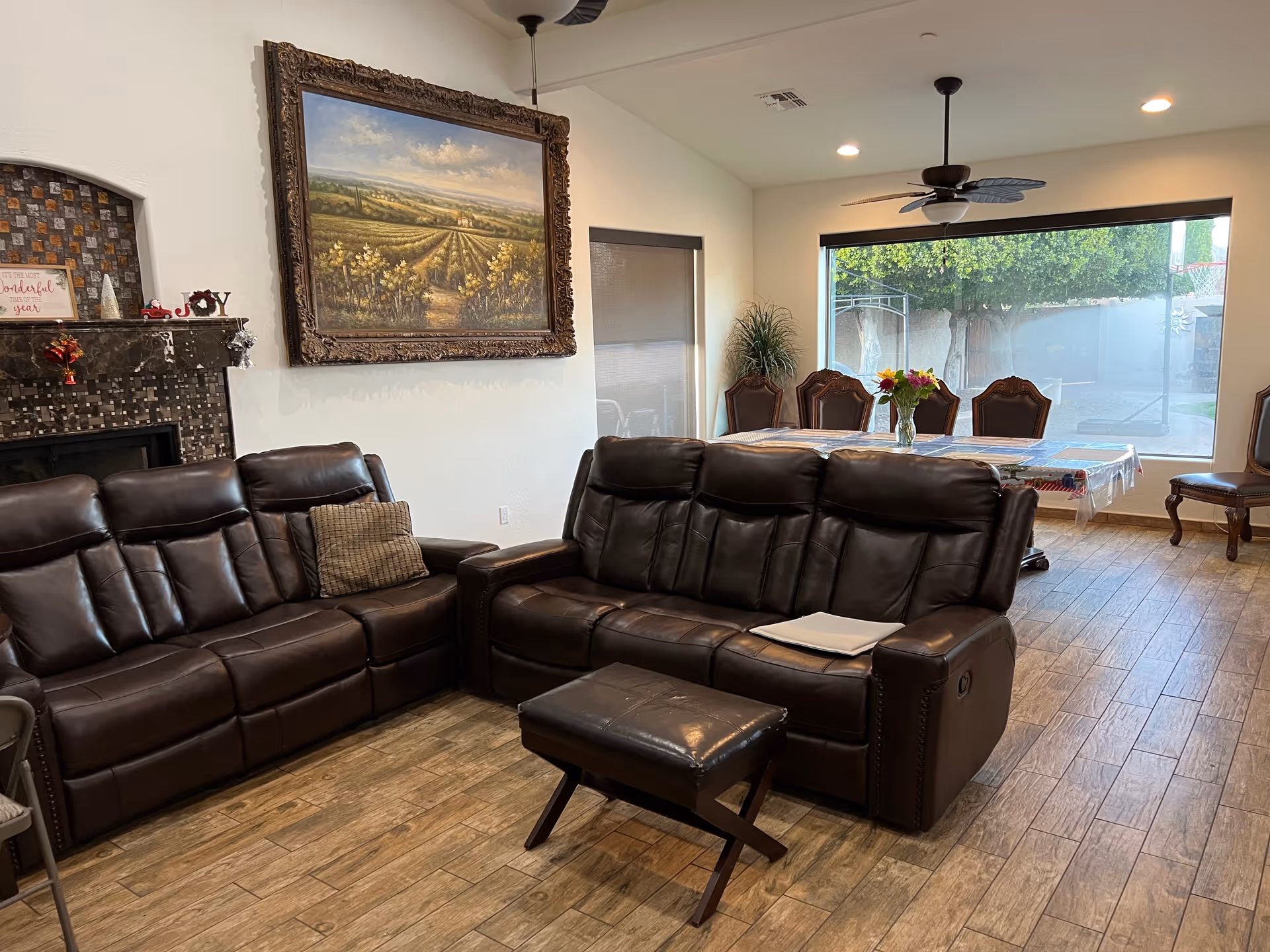Open living room and dining area with two brown leather reclining sofas, an ottoman, a fireplace, a large framed landscape painting, and a dining table by a big window.