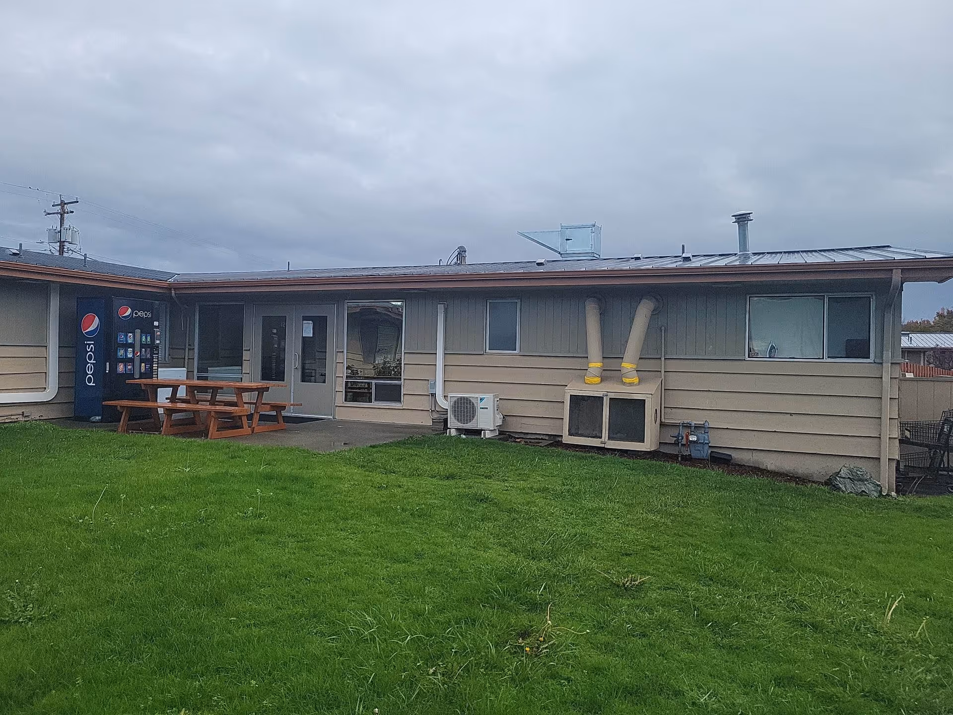 Outdoor view of a single-story building with beige siding and a metal roof under a cloudy sky. There is a green grassy area in the foreground, a wooden picnic table near the building, a Pepsi vending machine to the left, and two ventilation pipes attached to a unit on the wall. Windows and a glass door are visible on the building's exterior.
