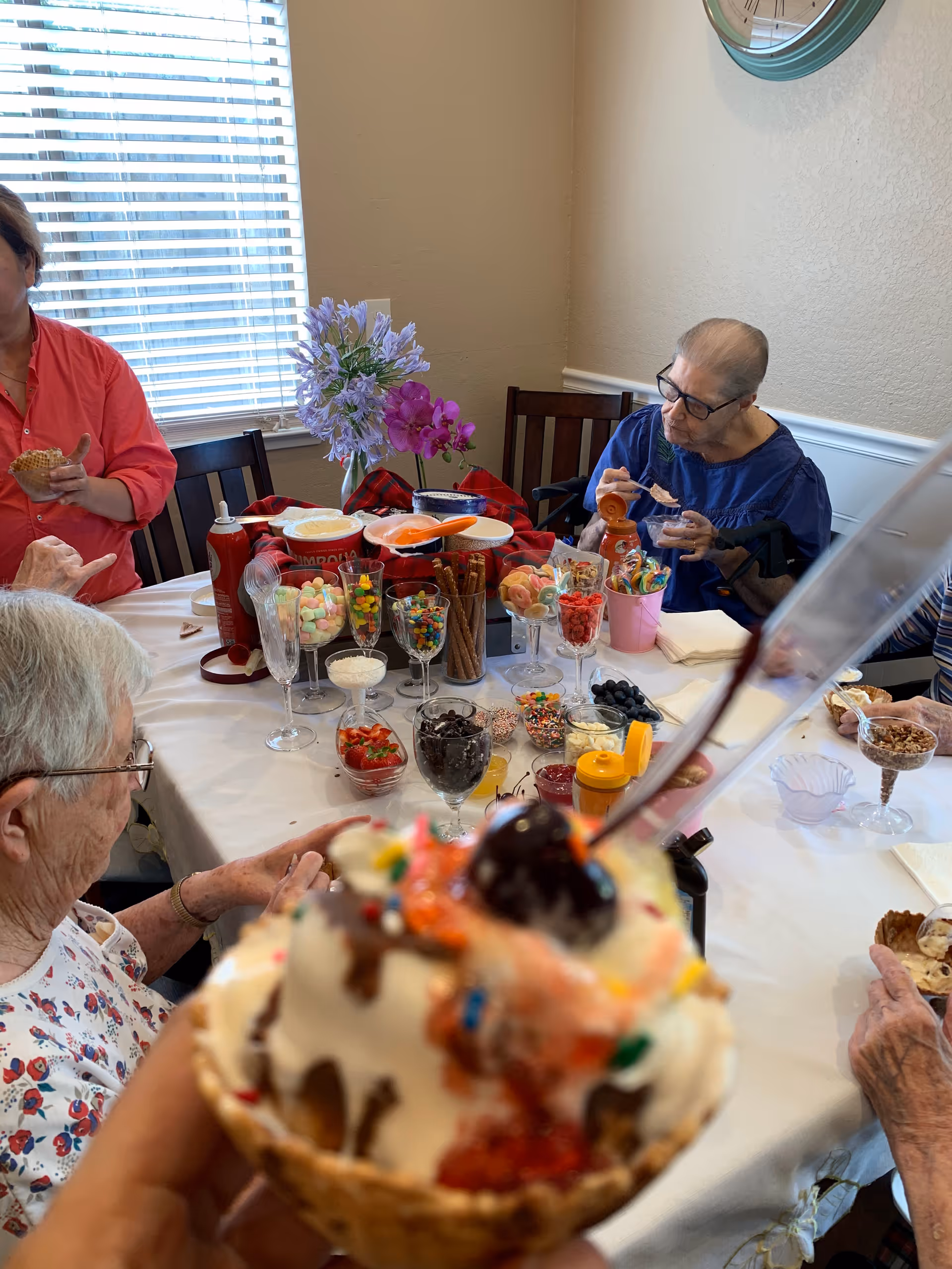 People gathered around a table with an ice cream sundae bar and assorted toppings, with a waffle-bowl sundae held in the foreground.