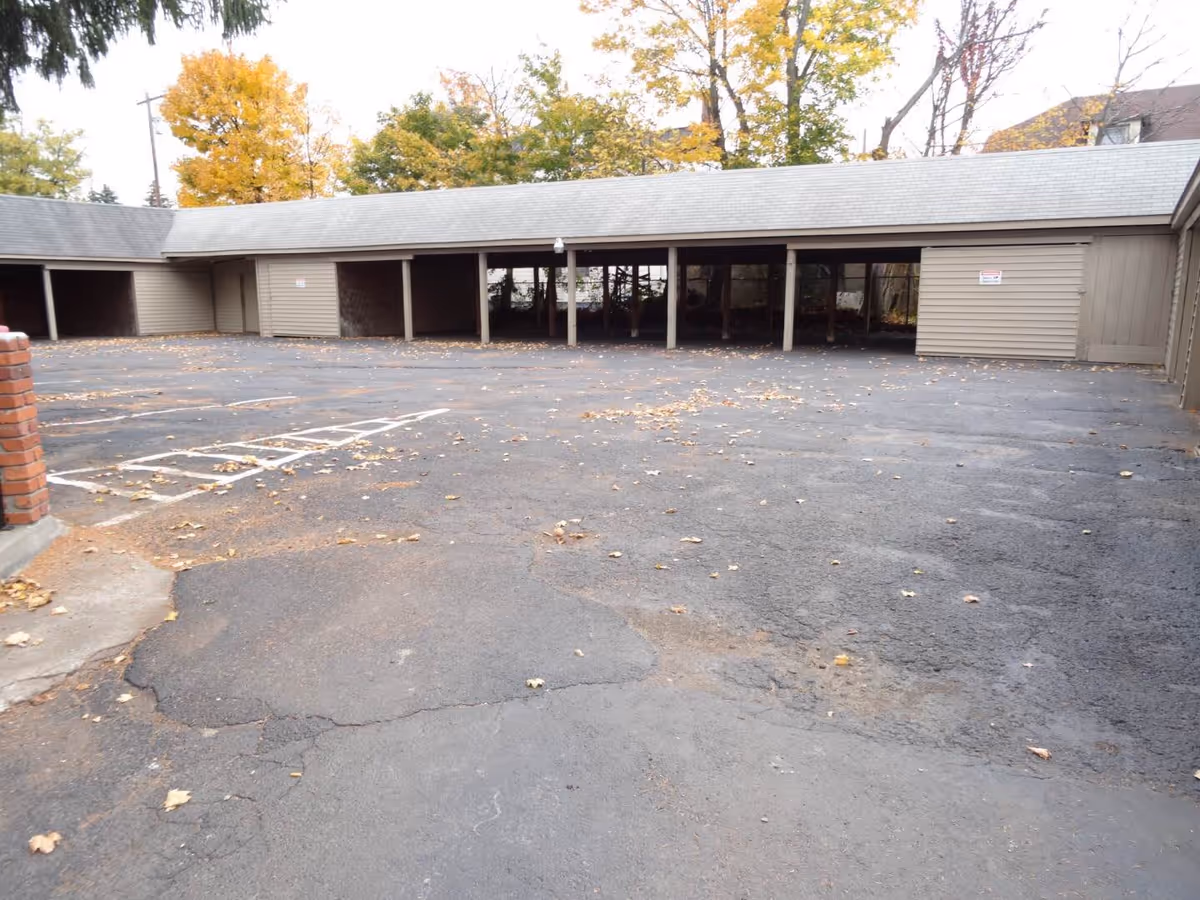 Outdoor parking area with covered parking spaces and a paved asphalt surface scattered with fallen leaves. Trees with autumn foliage are visible in the background.