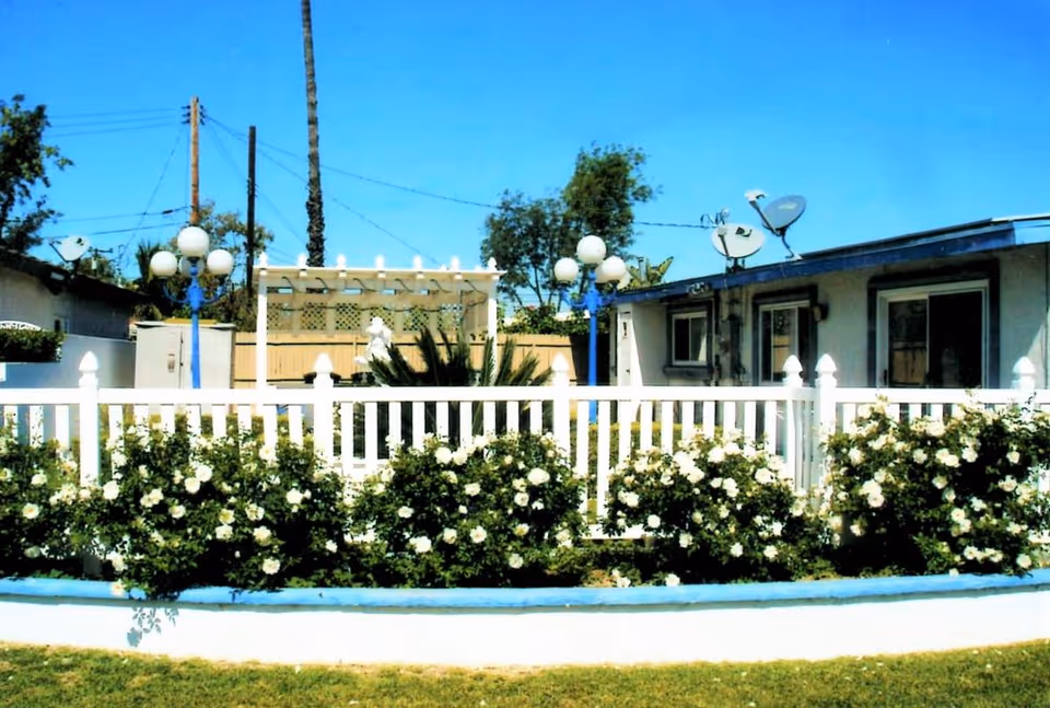 Outdoor view of a senior living facility with a white picket fence and blooming white flowers in front. The building has satellite dishes on the roof, and there are two lamp posts with multiple round lights. Trees and power lines are visible in the background under a clear blue sky.