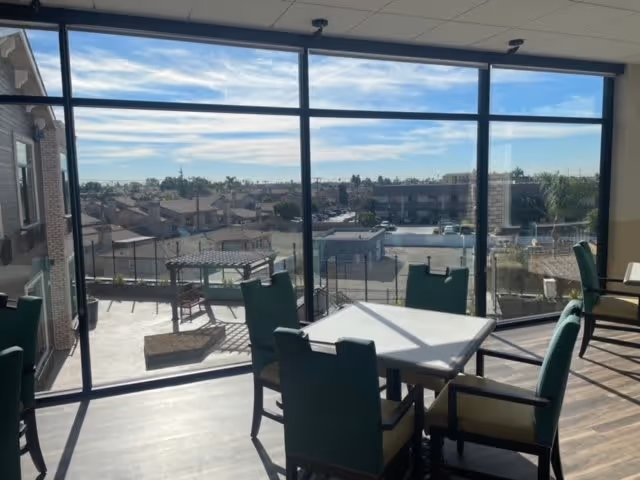 Interior view of a room with large floor-to-ceiling windows showing an outdoor patio and a neighborhood beyond. The room contains tables and chairs with green upholstery, and sunlight casts shadows on the floor.