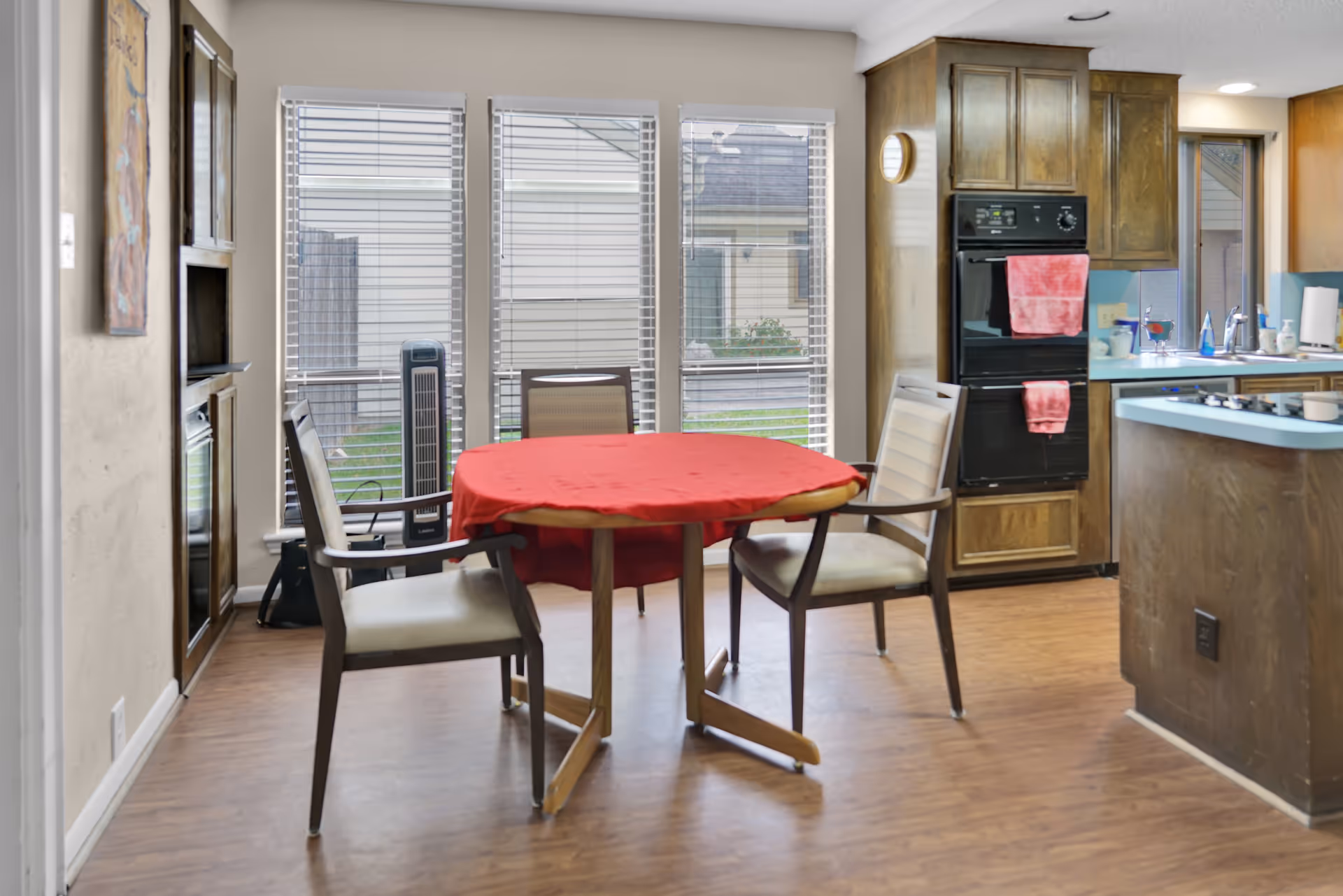 A dining area with a round table covered by a red tablecloth surrounded by four chairs. The room has large windows with blinds, a wooden floor, and a kitchen area with wooden cabinets, a double oven with red towels hanging, and a countertop with a sink and various items.