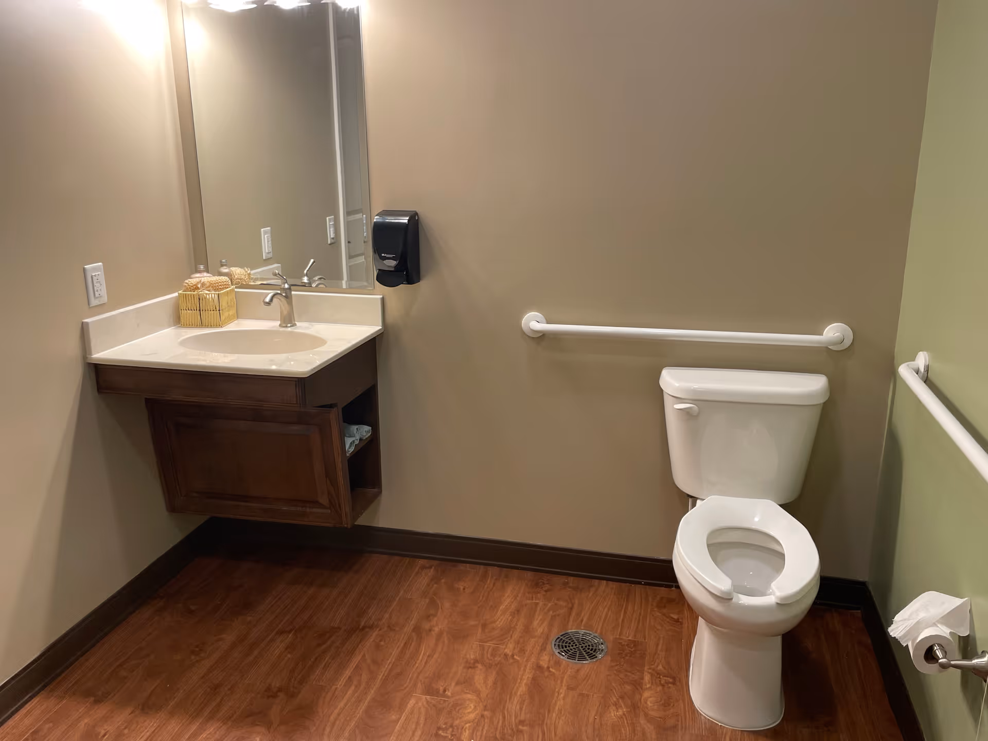 A clean and accessible bathroom with a white toilet, grab bars on the walls, a wall-mounted sink with a wooden cabinet underneath, a large mirror above the sink, a soap dispenser, and a tissue holder. The floor has a wood-like finish.