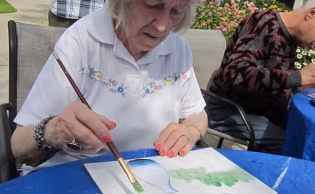 An elderly woman with white hair is sitting outdoors at a table covered with a blue tablecloth, painting on a piece of paper. She is holding a paintbrush and appears focused on her artwork. Another person is seated next to her, also engaged in an activity. There are flowers and greenery in the background.