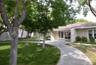 Curved walkway leads to a single-story building entrance surrounded by green trees.