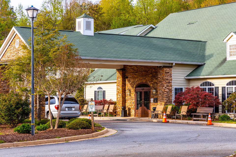 Entrance of Brasstown Manor Senior Living facility with a covered driveway, stone pillars, green roof, outdoor seating, and surrounding trees and shrubs.