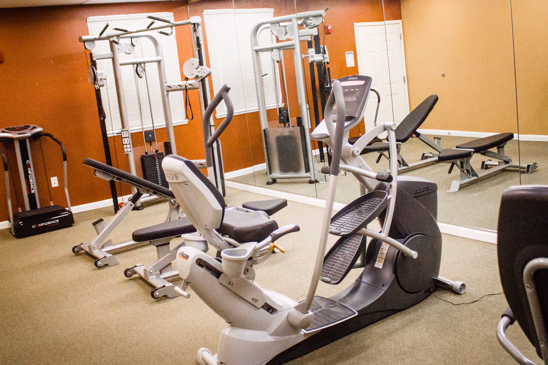 Indoor fitness room with cardio and weight machines, including an elliptical and benches in front of a mirrored wall.