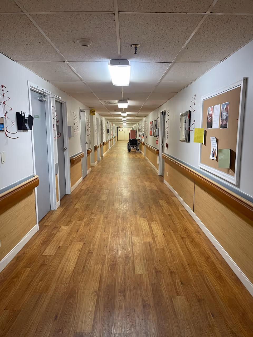 Long hallway in a skilled nursing and rehab facility with wood-patterned flooring, white walls with wooden paneling halfway up, multiple closed doors on both sides, ceiling lights, a bulletin board on the right wall, and a wheelchair positioned near the far end.