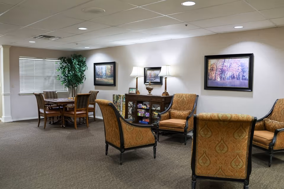 A cozy common area in Aberdeen Heights Assisted Living featuring four upholstered armchairs arranged around a small table, a wooden cabinet with board games, two table lamps, framed landscape paintings on the wall, a dining table with six chairs near a window with blinds, and a large potted plant in the corner.