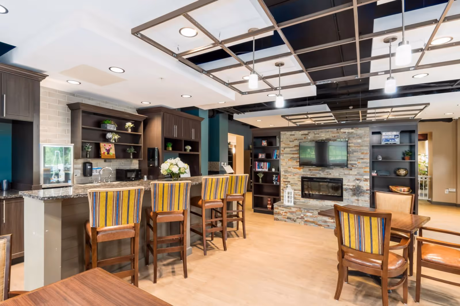 A bright communal lounge with a granite-topped bar, striped bar stools, dining tables, shelving, and a stone fireplace with a wall-mounted TV.