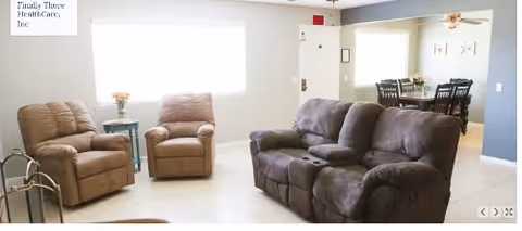 A bright living room with two beige recliner chairs and a dark gray reclining sofa facing a large window. A small side table with a flower vase is placed between the recliners. In the background, there is a dining area with a table and four chairs, and a ceiling fan above it.