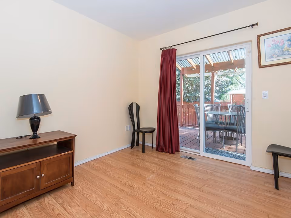A room with light beige walls and wooden flooring featuring a wooden cabinet with a black lamp on top, two black chairs, and a sliding glass door with red curtains leading to an outdoor patio with a table and chairs under a pergola.